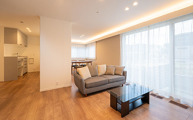 【A区画リビング】20畳以上の広々としたLDKはインテリアコーディネーターおすすめのアクセントクロスと窓上のコーブ照明でおしゃれな空間に。ソファとテレビボード付きです。（2025年5月撮影）