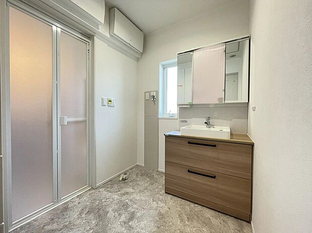 【洗面・脱衣所】広々とした脱衣室は、木目調の洗面台が温かみを添える清潔感のある空間。ゆとりある広さを確保しており、忙しい朝の準備もスムーズです。使い勝手の良い収納と明るい照明で、日々の身支度を快適にサポートします。