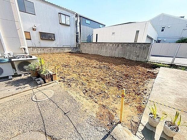 【着工前現地画像】整地された更地の外観写真です。奥に擁壁と隣家があり、右側に敷地入口の舗装。道路幅約4mのアスファルト道路に面し、土地面積約91.28m2です。