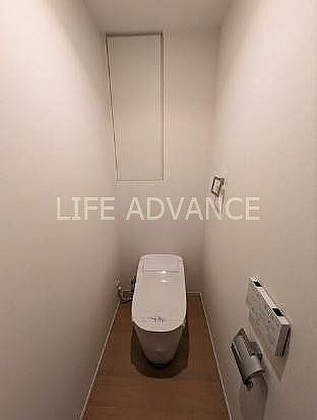 白を基調としたシンプルなトイレには、温水洗浄便座や壁掛け式のリモコン、ペーパーホルダーが備え付けられており、正面奥の壁面には扉付きの埋め込み収納棚が設置されております。