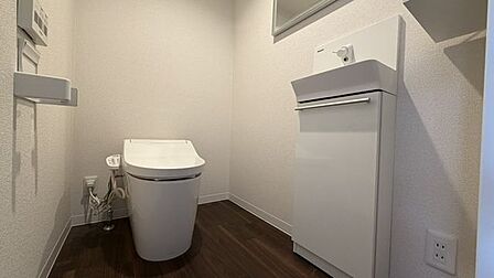 白を基調とした壁紙に落ち着いた木目調の床を合わせたトイレには、温水洗浄機能付きの便座と手洗い器が設置されており、手洗器の下部には収納スペースを備えています。