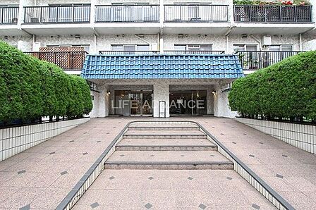 青い屋根が特徴的なエントランスアプローチには、中央の階段に加えて両脇にゆるやかなスロープが設けられており、アプローチの左右には整えられた生垣とタイル貼りの床面が広がっています。