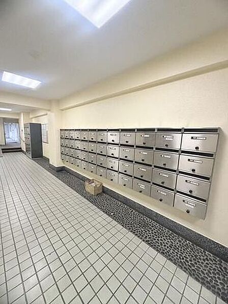 共用部の壁面にはダイヤル錠を備えたステンレス製の集合ポストが整然と設置されており、床面は白の格子状タイル貼りで、奥にはロッカー設備や通路が続いています。