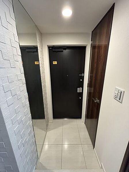 光沢感のあるタイル敷きの玄関スペースには、壁面に大型の姿見鏡が設置されており、立体感のある凹凸デザインの壁材や黒調の玄関ドア、深みのある木目調の建具が採用されています。
