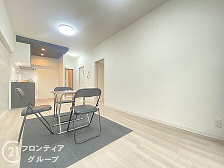 明るい木目調のフローリングとホワイトのクロスで統一された、広々としたLDK空間です。奥側にはキッチンが配置されています。