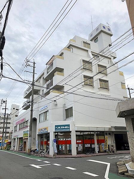 街角に佇む、白を基調とした特徴的なデザインの建物です。店舗と住居が一体となった複合施設で、周辺は電線が張り巡らされ、生活感のある環境がうかがえます。