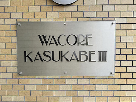 エントランスに掲げられたマンションの銘板です。レンガ調タイルの壁面に落ち着いた雰囲気で設置されており、住まいの顔となる佇まいです。※AIによる家具消し加工を行なっております。