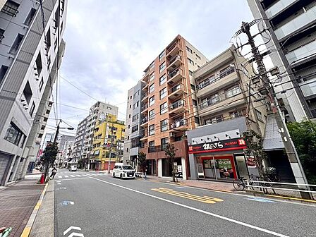 周辺に中高層の建物が立ち並ぶ街並みに調和したマンションです。前面道路は舗装され、建物沿いには歩道も整えられており、駅周辺の利便性を感じられる立地です。