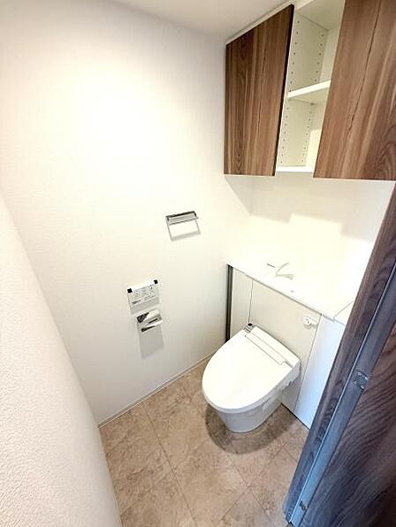 清潔感のあるトイレ空間です。収納棚が設置されており整理整頓がしやすい仕様です。明るい色合いで開放感を感じられます。温水洗浄便座も付いています。ドアや棚などの木目調がアクセントとなっています。