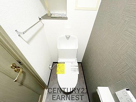 シンプルなデザインで清潔感を感じるトイレです。ペーパーホルダーや棚が設置され使い勝手も良好です。壁面のクロスと床材が調和した空間となっています。収納スペースも備えられており機能面も配慮されています。