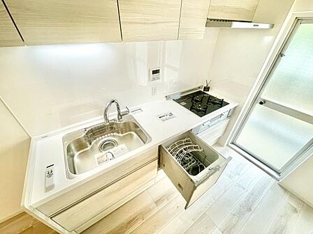 シンプルなデザインのキッチンは白を基調とし清潔感があります。食洗機や三口コンロなど設備も整っています。収納スペースも充実しており使い勝手の良さが感じられます。窓からの自然光が入りやすく明るい空間です。