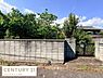 外観：現地土地写真には緑豊かな植栽としっかりとしたコンクリート塀が写されています。落ち着いた住宅街の雰囲気が感じられる一枚です。道路に面した場所に入り口と門扉が設けられています。